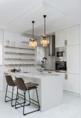A modern white kitchen with a speckled stone island, two dark brown bar stools, gold crystal pendants, and floating shelves.