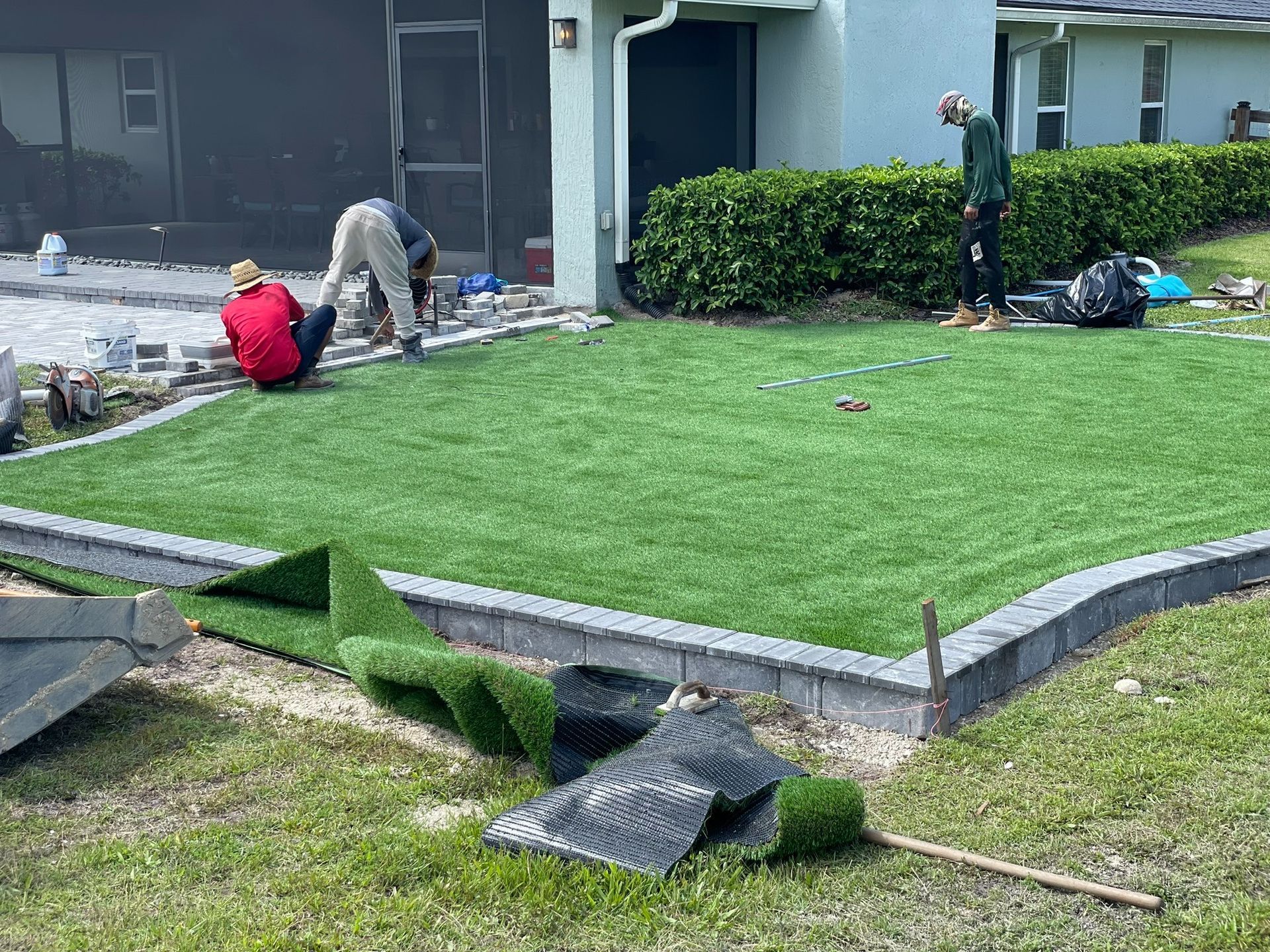 Workers installing sod on a front lawn beside a house, with rolled turf and tools nearby.