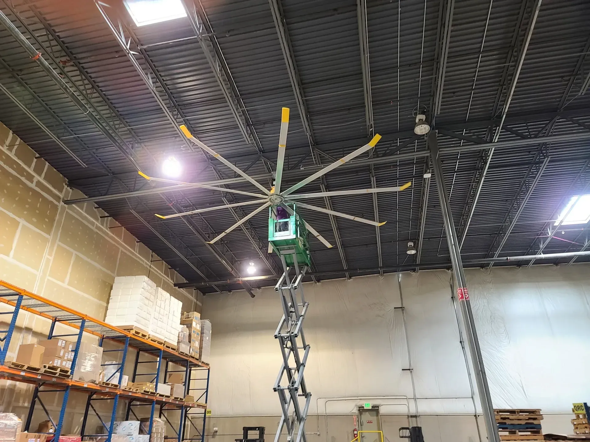 A large fan is hanging from the ceiling of a warehouse installed by B & W Services Technician.