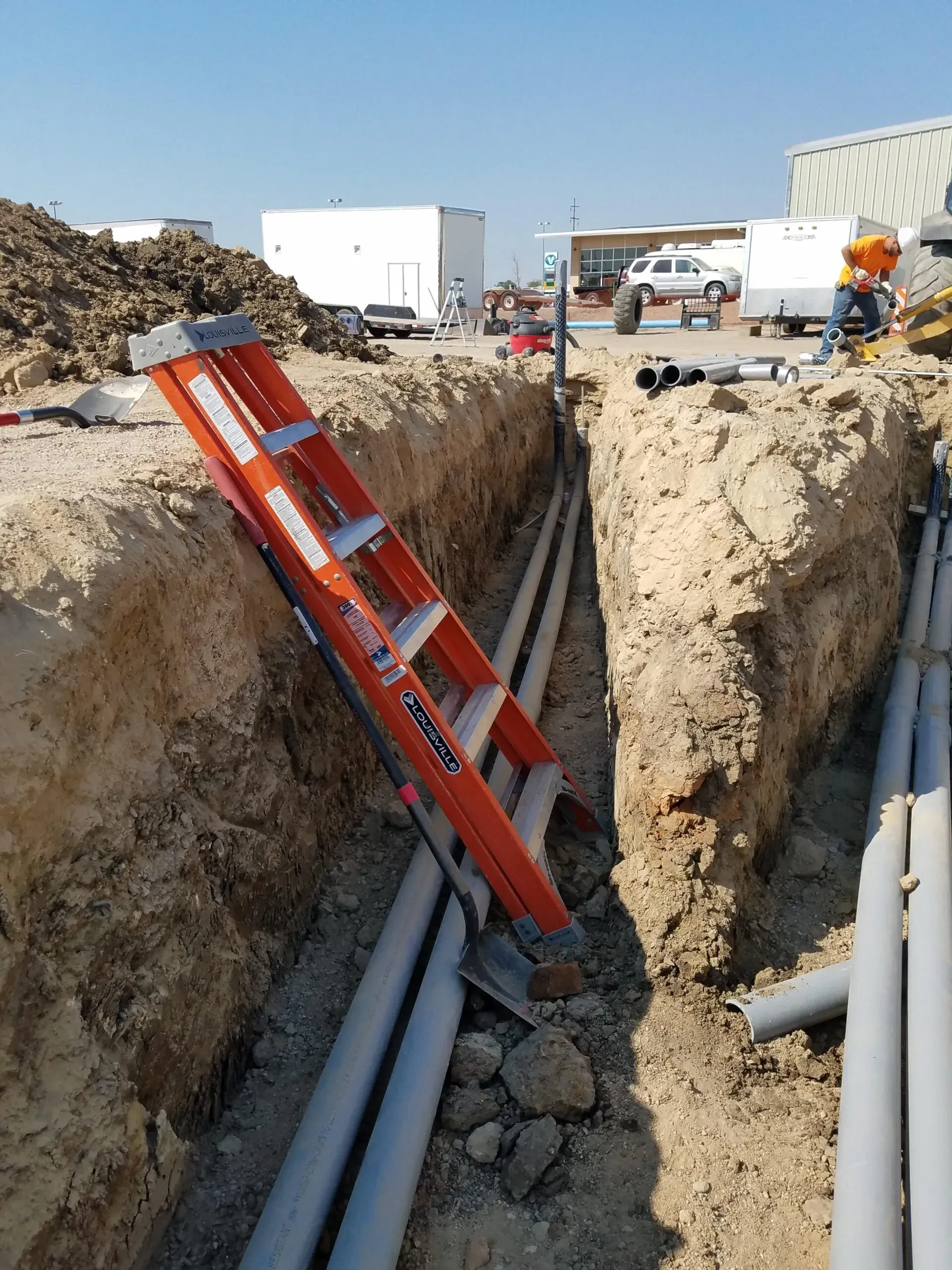 A ladder is laying in the dirt next to a trench filled with pipes.