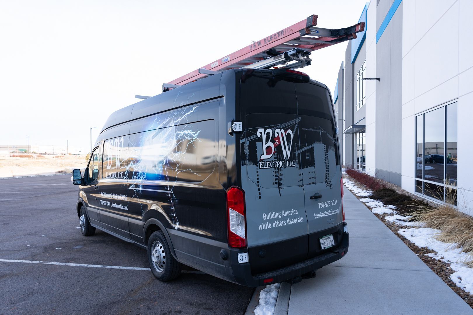 A black B & W Services van with a ladder on top of it is parked in front of a building.