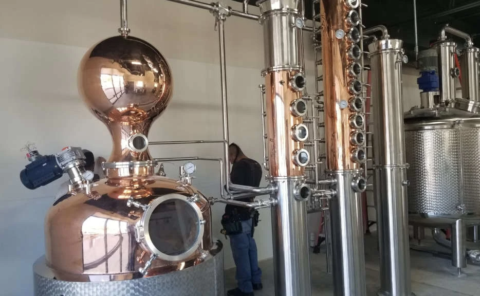 A man is standing in front of a large copper distillery.