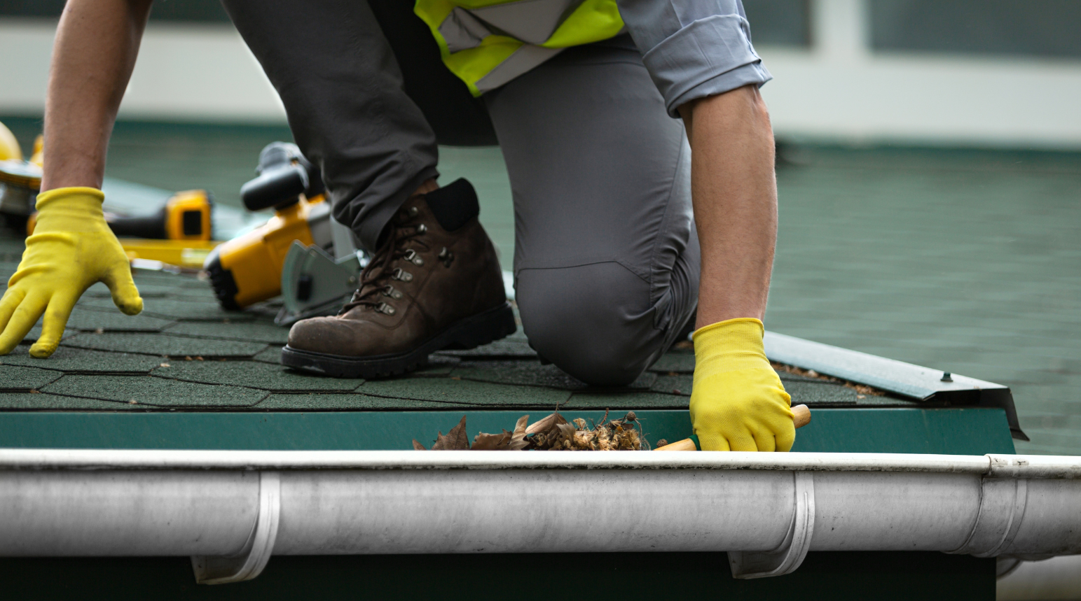 Construction worker in yellow gloves and work boots cleaning a roof gutter.