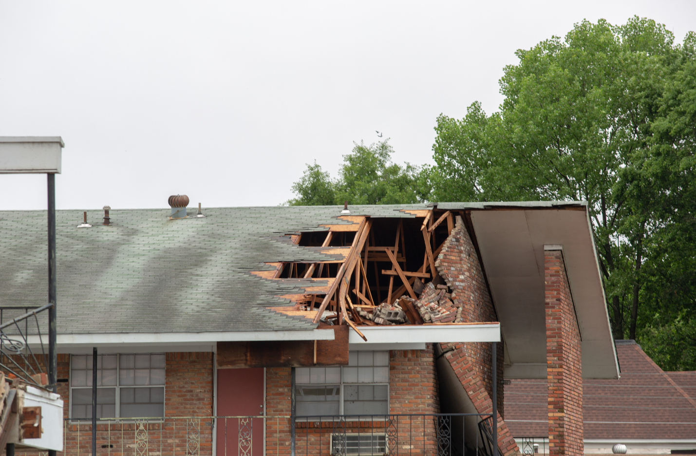 Damaged roof on a brick apartment building, revealing wooden supports. Overcast sky with green trees in the background.