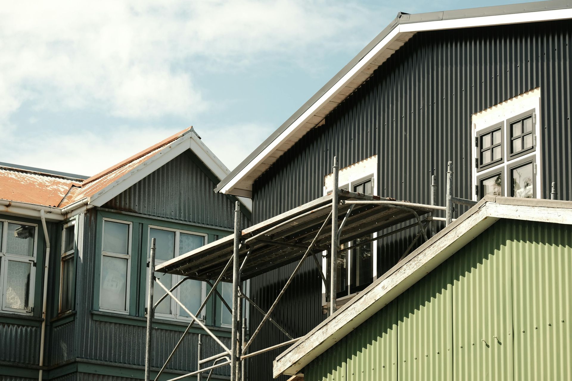 Buildings with corrugated metal siding in shades of green and gray, under a cloudy sky, with scaffolding present.