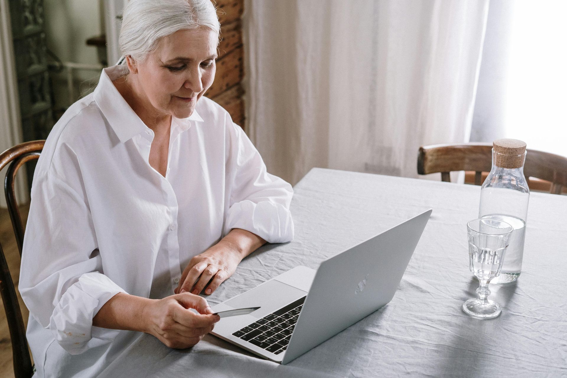 An older woman with gray hair sits at a table using a laptop, holding a credit card. 