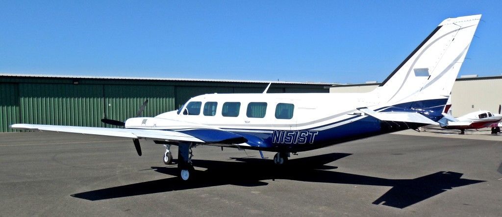 A white and blue airplane parked on an asphalt surface. In the background is a green wall and a blue sky.