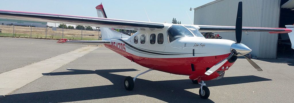 A red and white airplane on a runway. A propeller faces the viewer. A building is in the background.