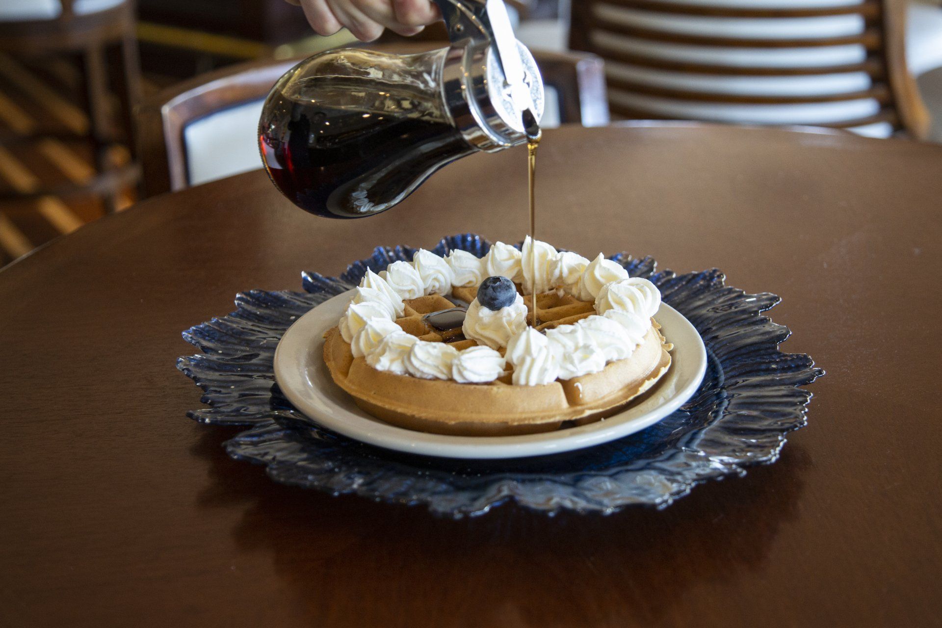 A person is pouring syrup on a waffle with whipped cream and blueberries.