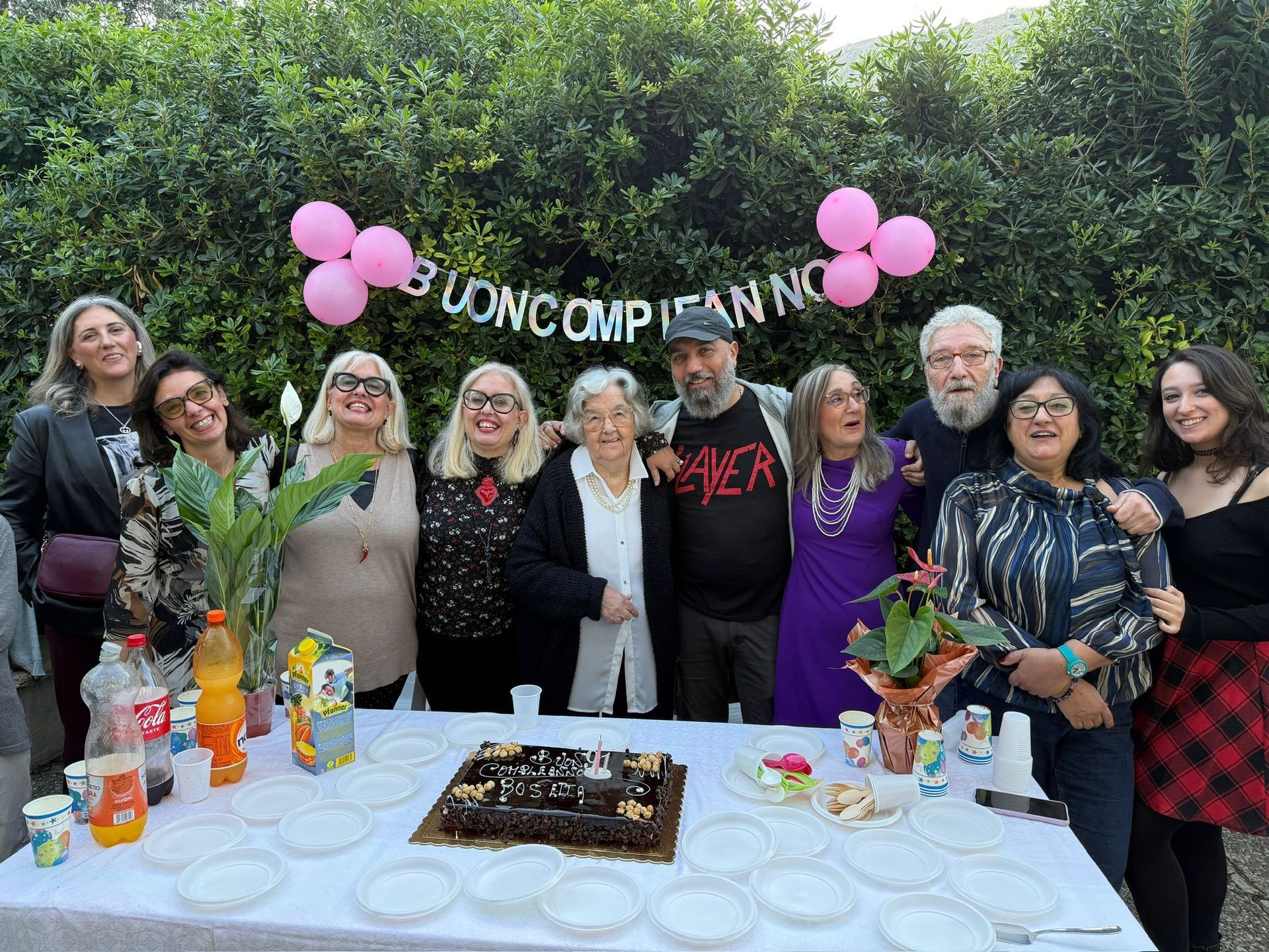 Il gruppo si è riunito attorno a un tavolo con torta e bevande, festeggiando all'aperto sotto palloncini rosa e uno striscione con la scritta