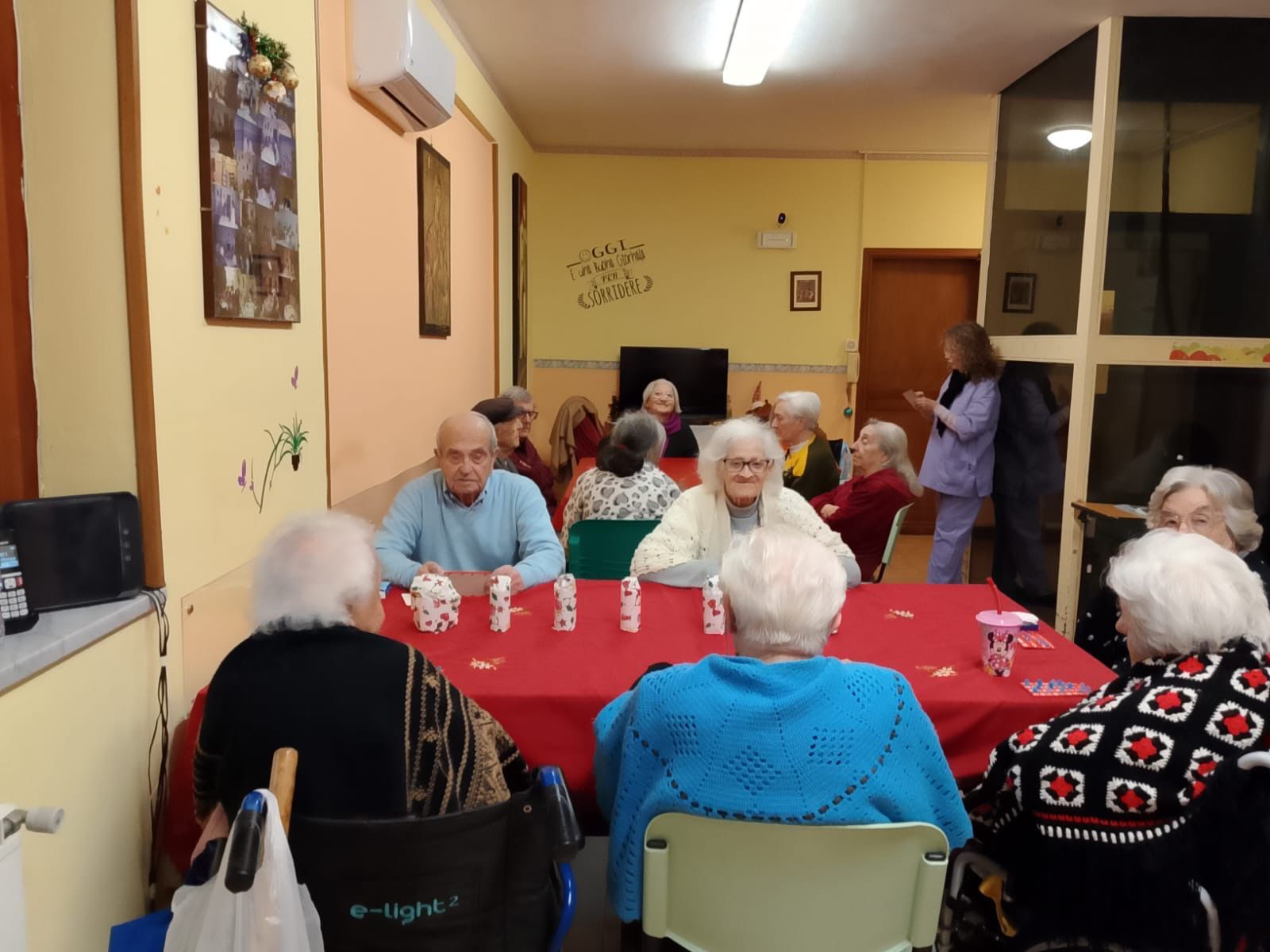 In una sala comune, alcune persone sono sedute attorno a una tovaglia rossa e chiacchierano durante un incontro di gruppo.
