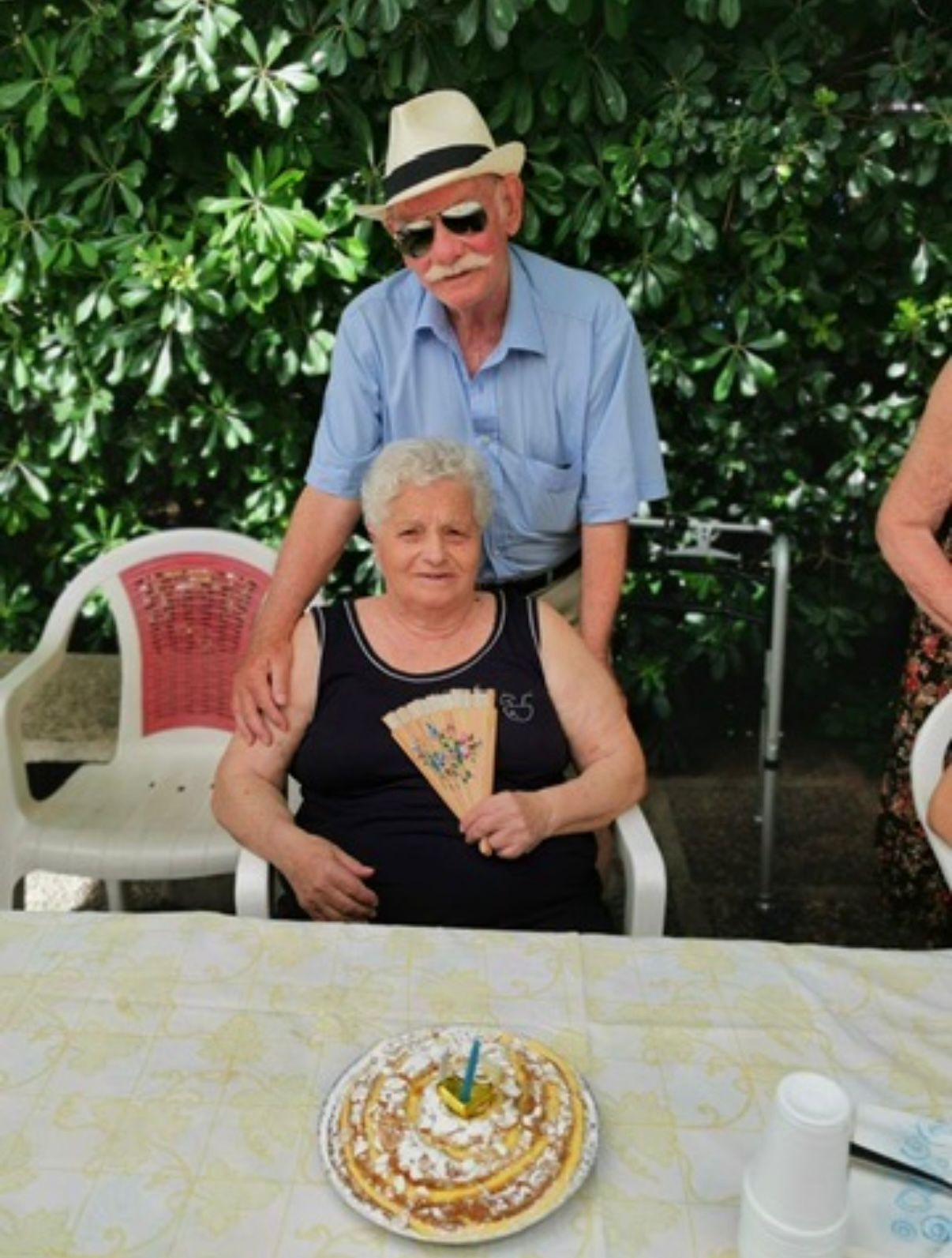 Due persone sedute a un tavolo all'aperto con una torta davanti a loro, circondate dal verde.