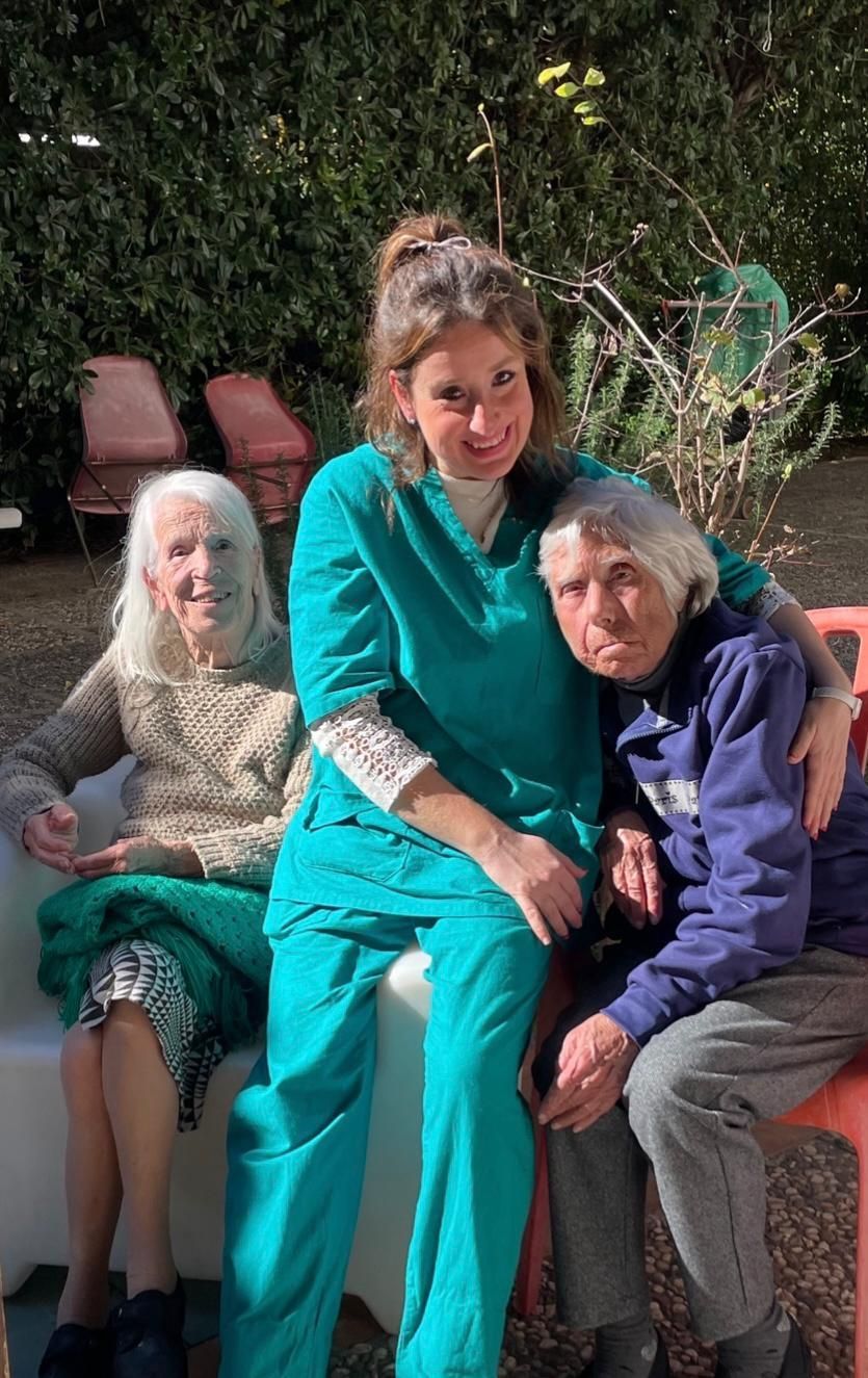 Tre persone sedute su una panchina all'aperto, con la persona al centro vestita di color verde acqua che sorride e abbraccia le altre.