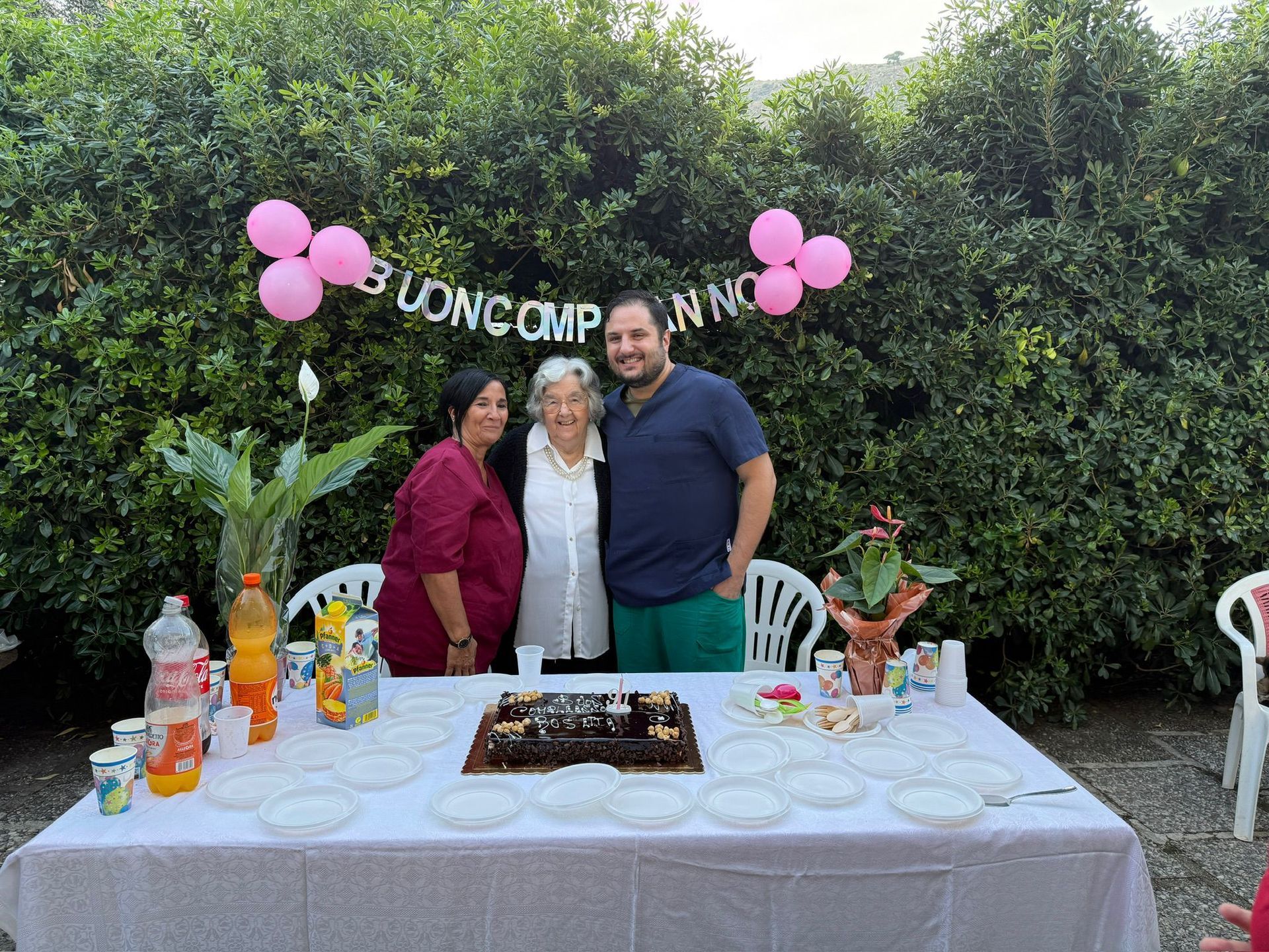 Persone in posa dietro un tavolo di compleanno all'aperto con palloncini rosa e uno striscione con la scritta