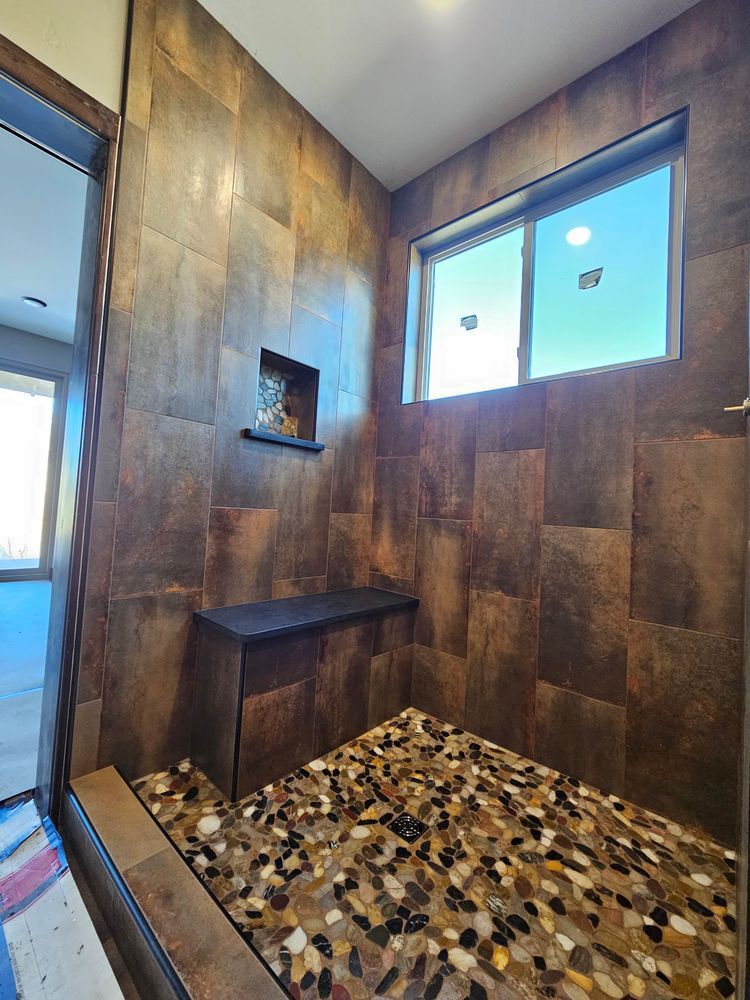 A bathroom with brown tile walls, pebble floor, bench, and a small window.