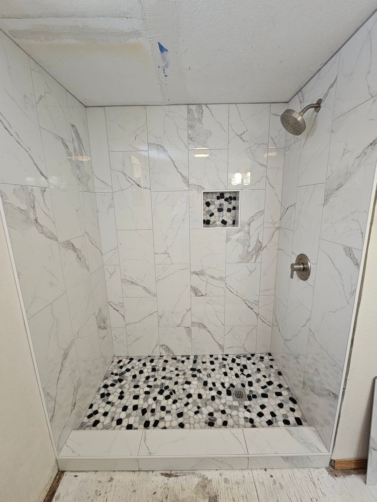 Newly tiled shower with marble-look walls, pebble floor, and a small inset shelf.