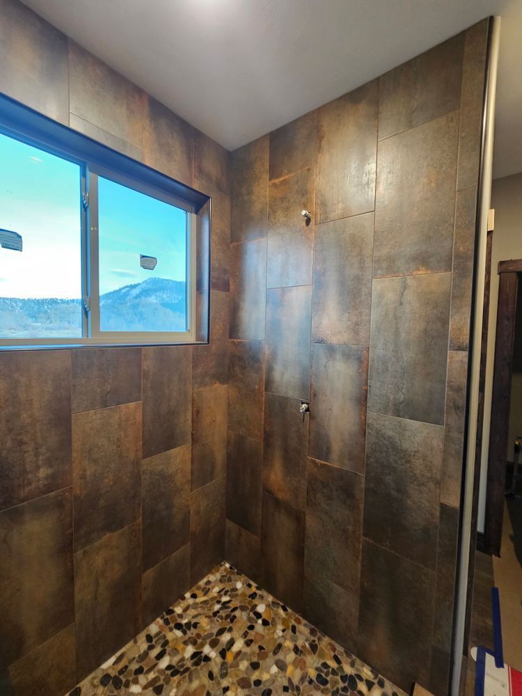 Shower with brown-toned tile walls and a pebble floor, beside a window.