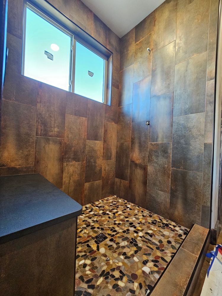 Shower with brown stone tile walls and pebble floor. Window above a built-in bench.