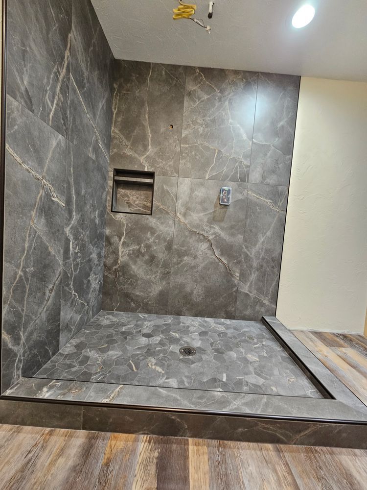 Gray tiled shower with recessed shelf, drain, and raised curb, on wood-look flooring.