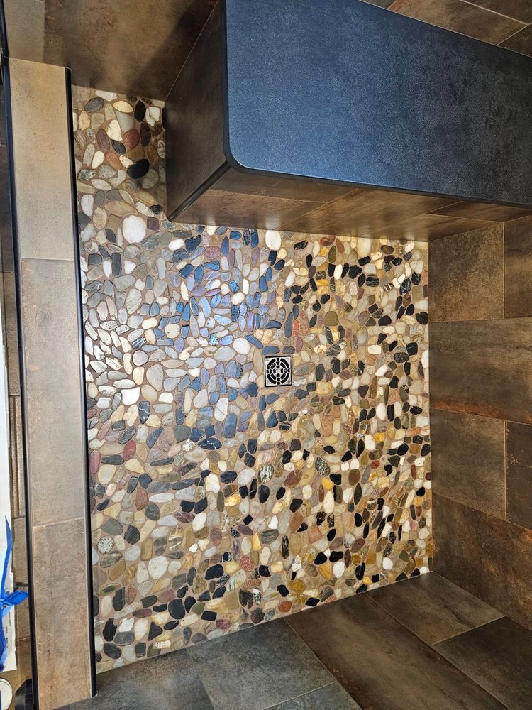 Shower floor with river rock tile, a dark drain, and wood-look walls.