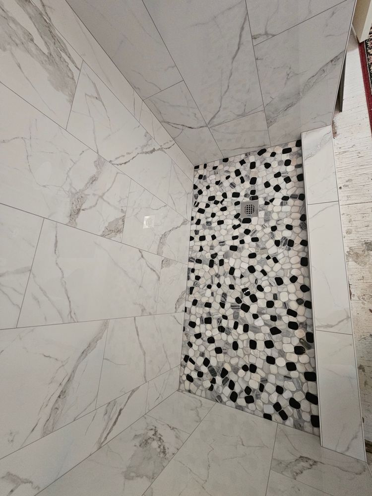 Shower interior with large white marble-like tiles on walls, small black/white mosaic tile floor, and shower head.