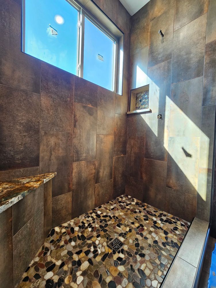Shower with pebble floor and dark brown tiled walls, with a window and shelf.