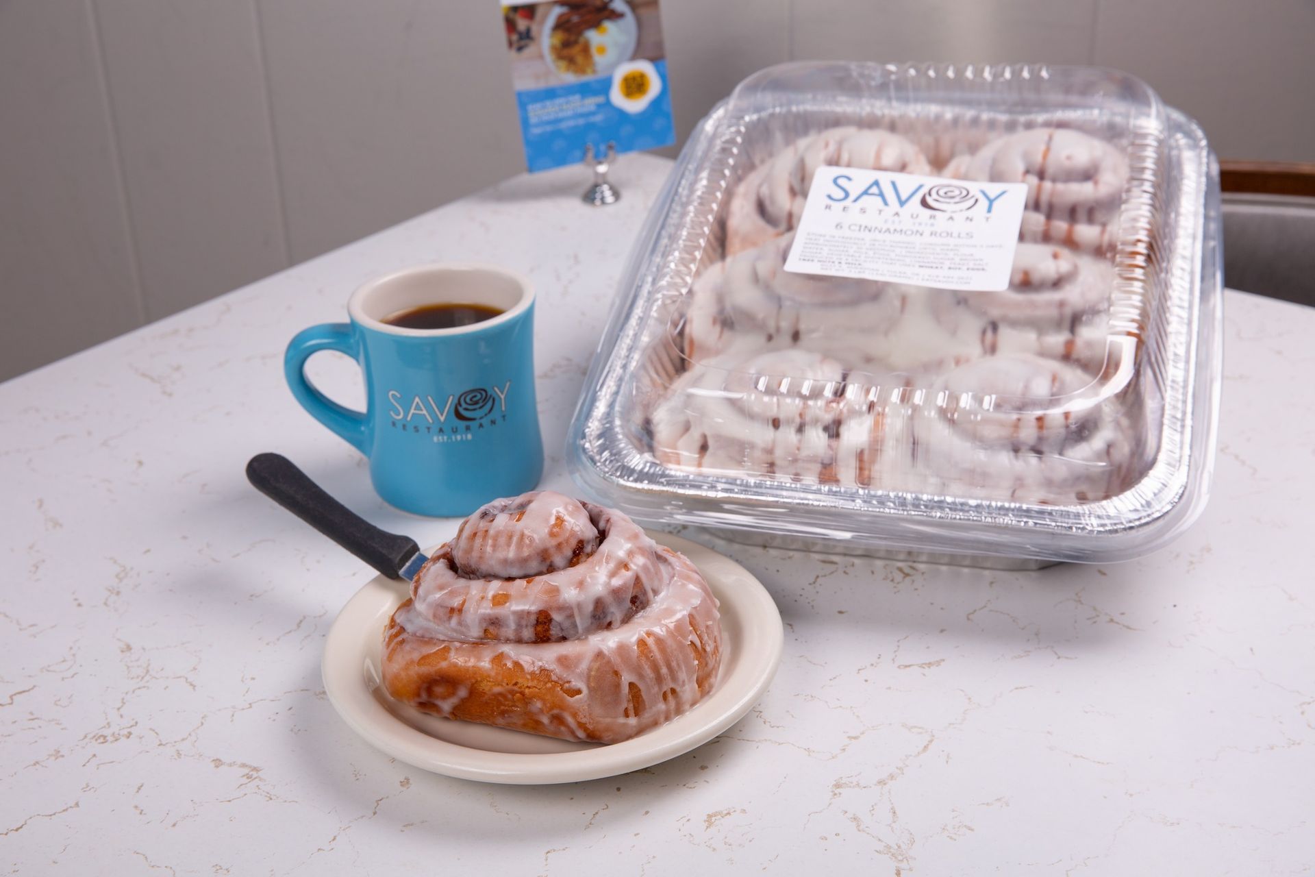 A tray of cinnamon rolls is on a table next to a plate of cinnamon rolls and a cup of coffee.