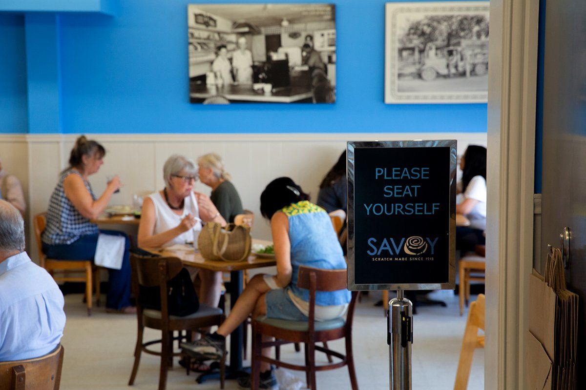 People are sitting at tables in a restaurant with a sign that says please seat yourself