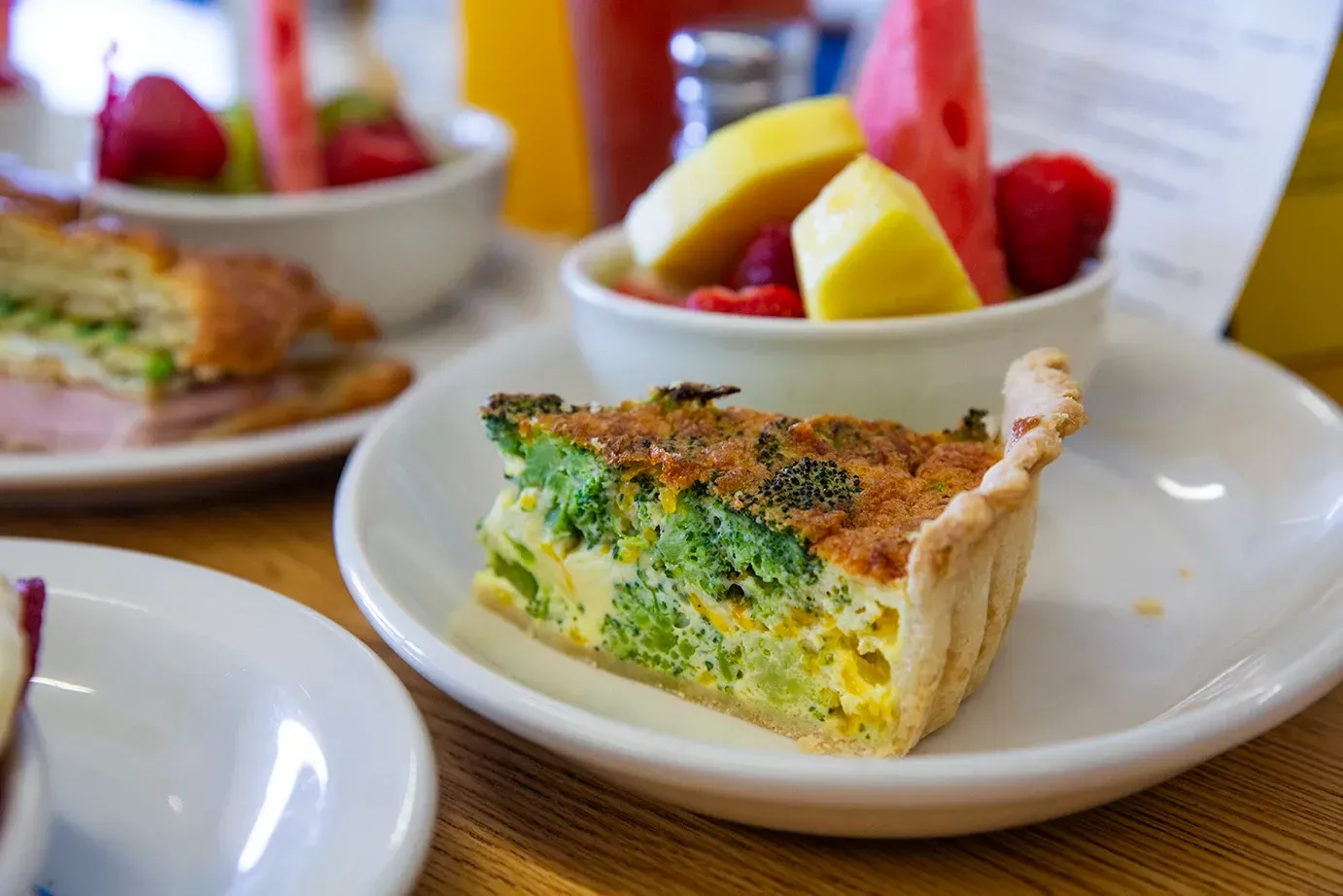 A slice of quiche is on a plate next to a bowl of fruit.
