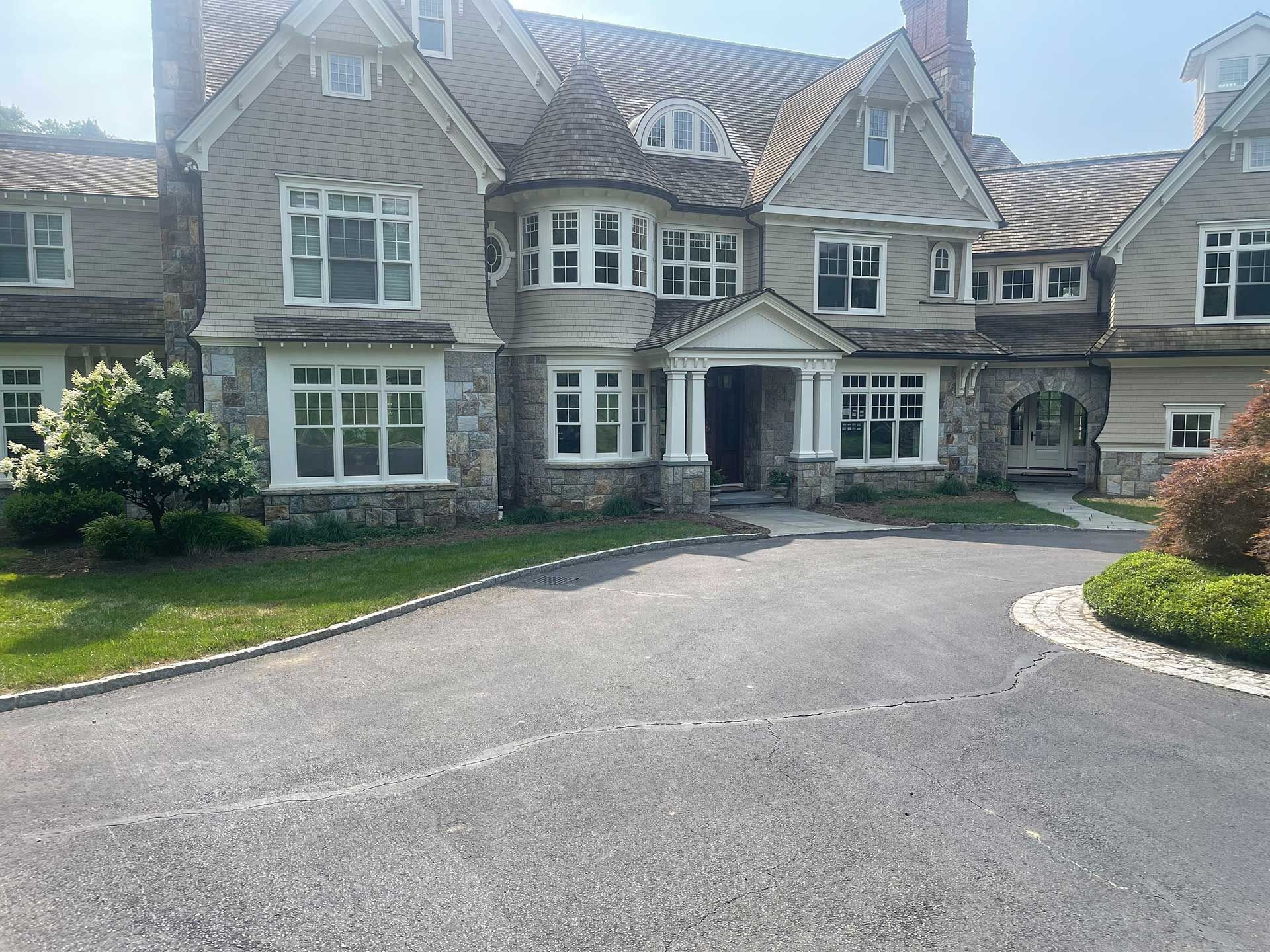 Large stone mansion with arched entry, manicured lawn, and curved driveway under a bright sky