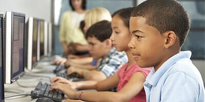 Children in a computer lab, using computers with a teacher in the background.