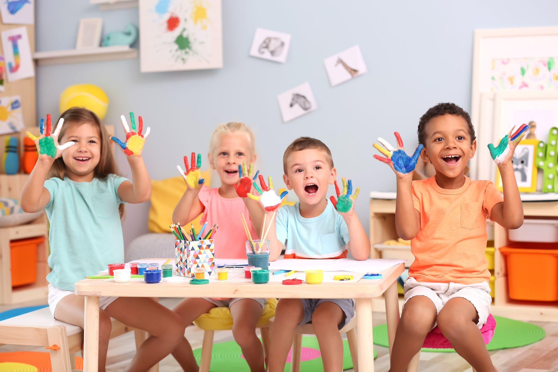 four children sitting at a table in a colorful classroom, showing their hands covered in bright 