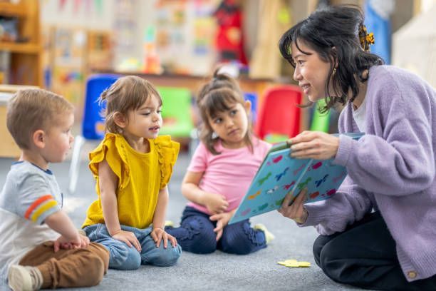 A preschool teacher sits on the floor of her classroom with a small group of students as she reads t