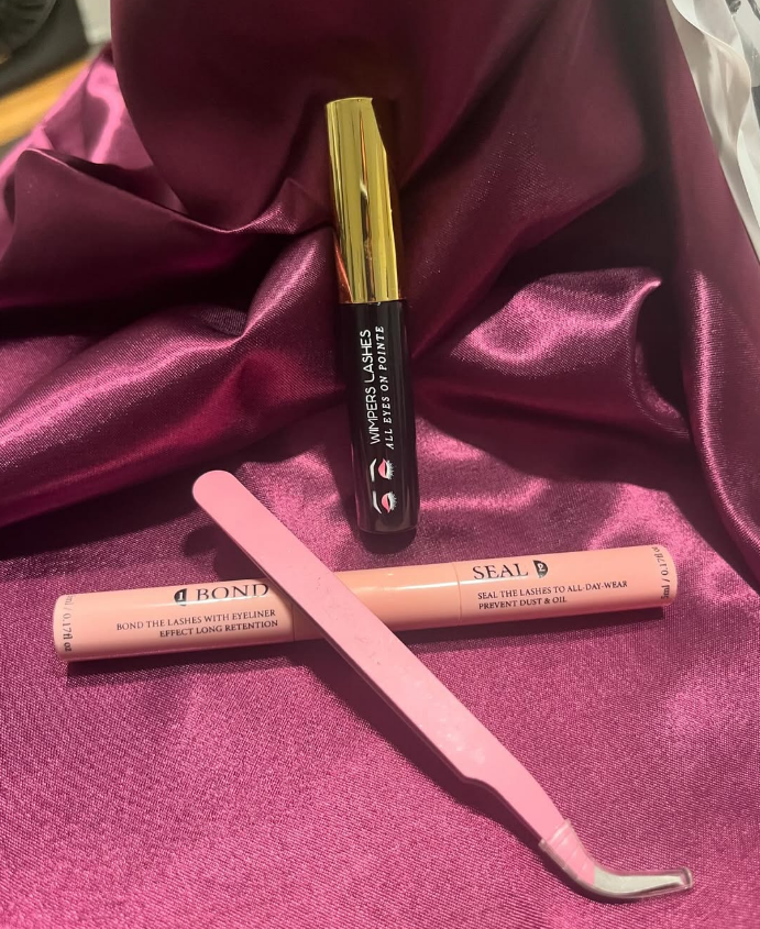 Pink-handled tweezers, pink eyeliner, and black mascara with gold lid on magenta satin fabric.