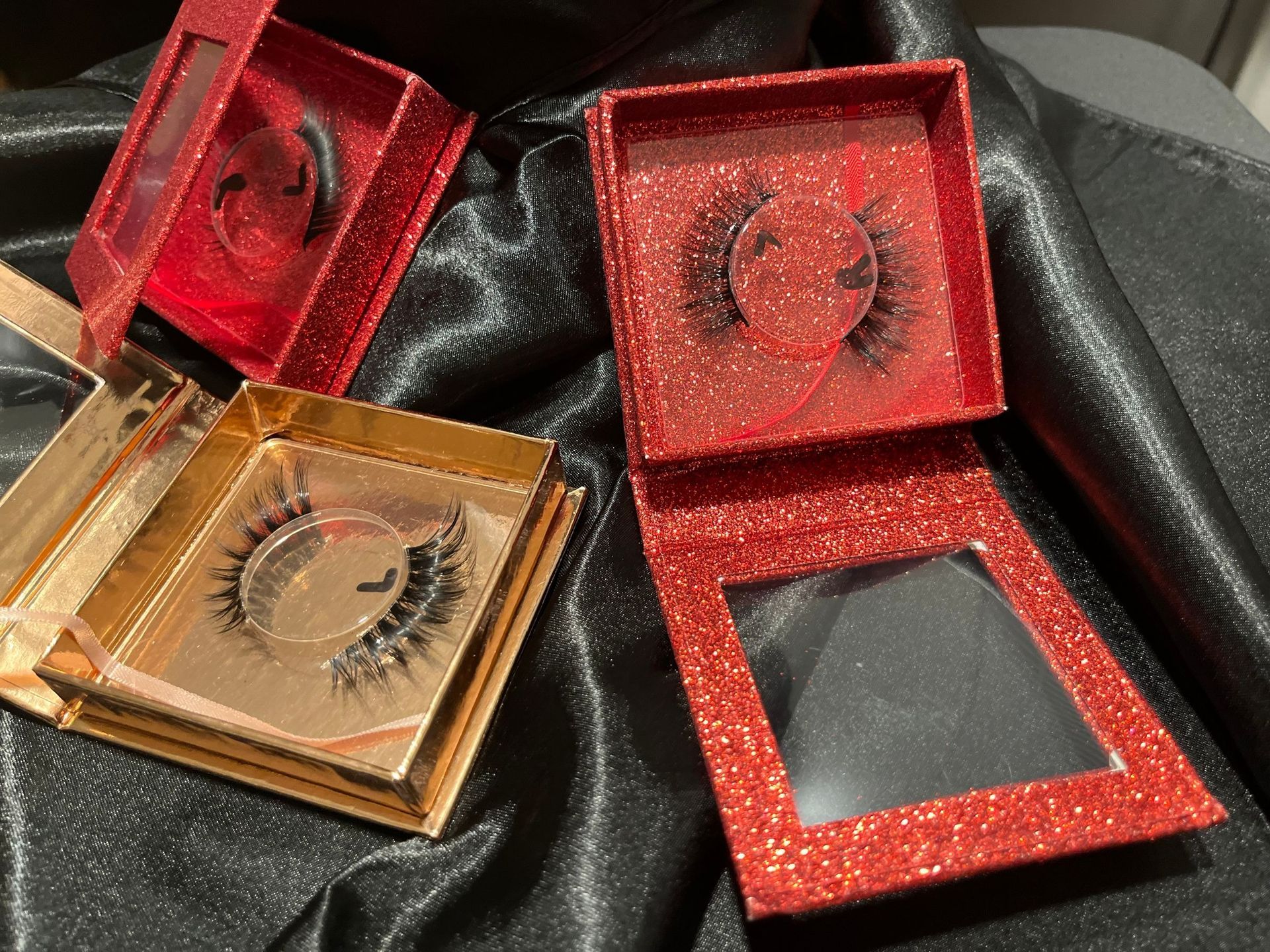 Red and gold glitter eyelash cases with false eyelashes against a black fabric background.