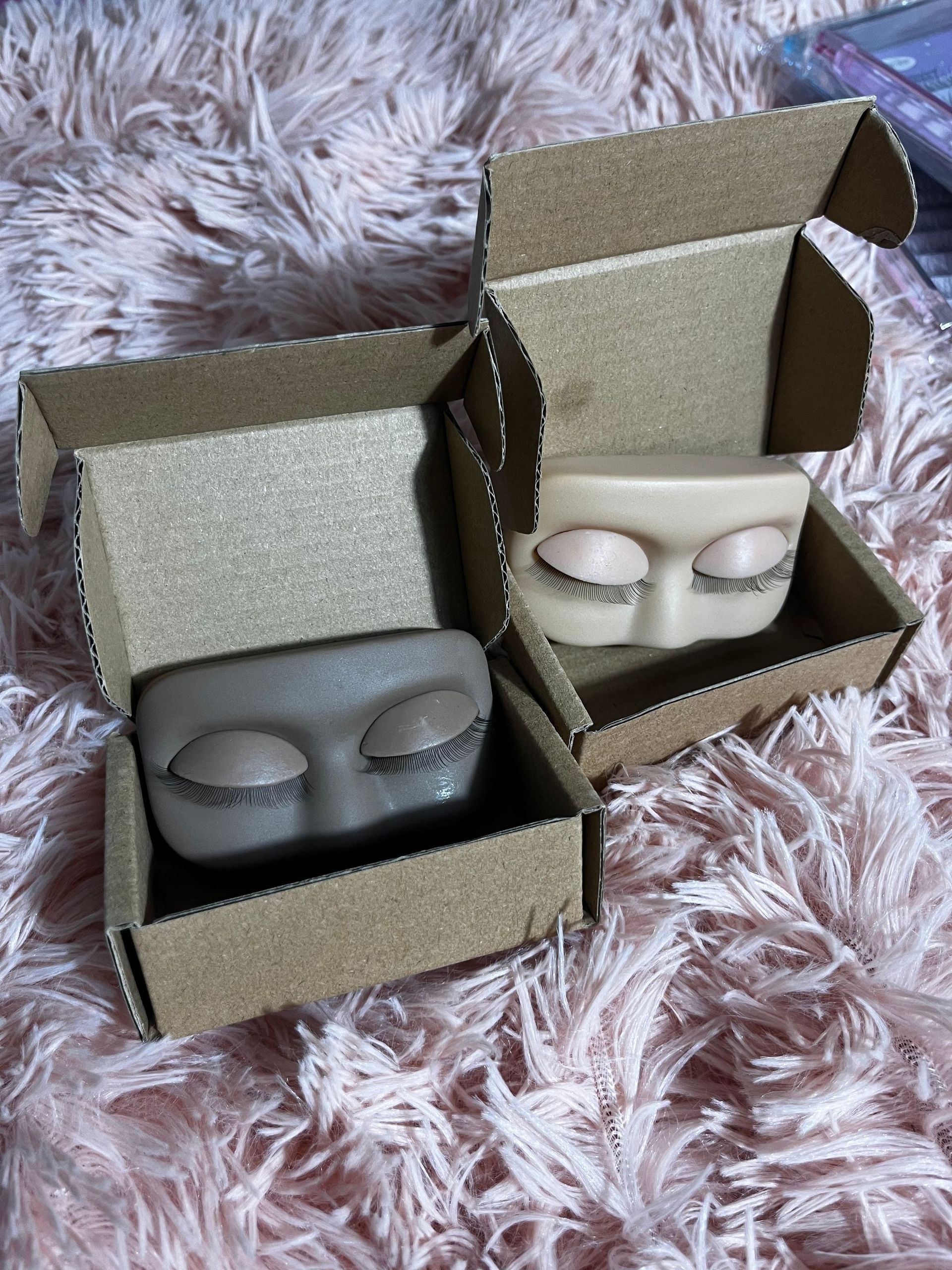 Two face-shaped, closed-eye soap bars inside open cardboard boxes on a pink furry surface. One is gray, one is beige.