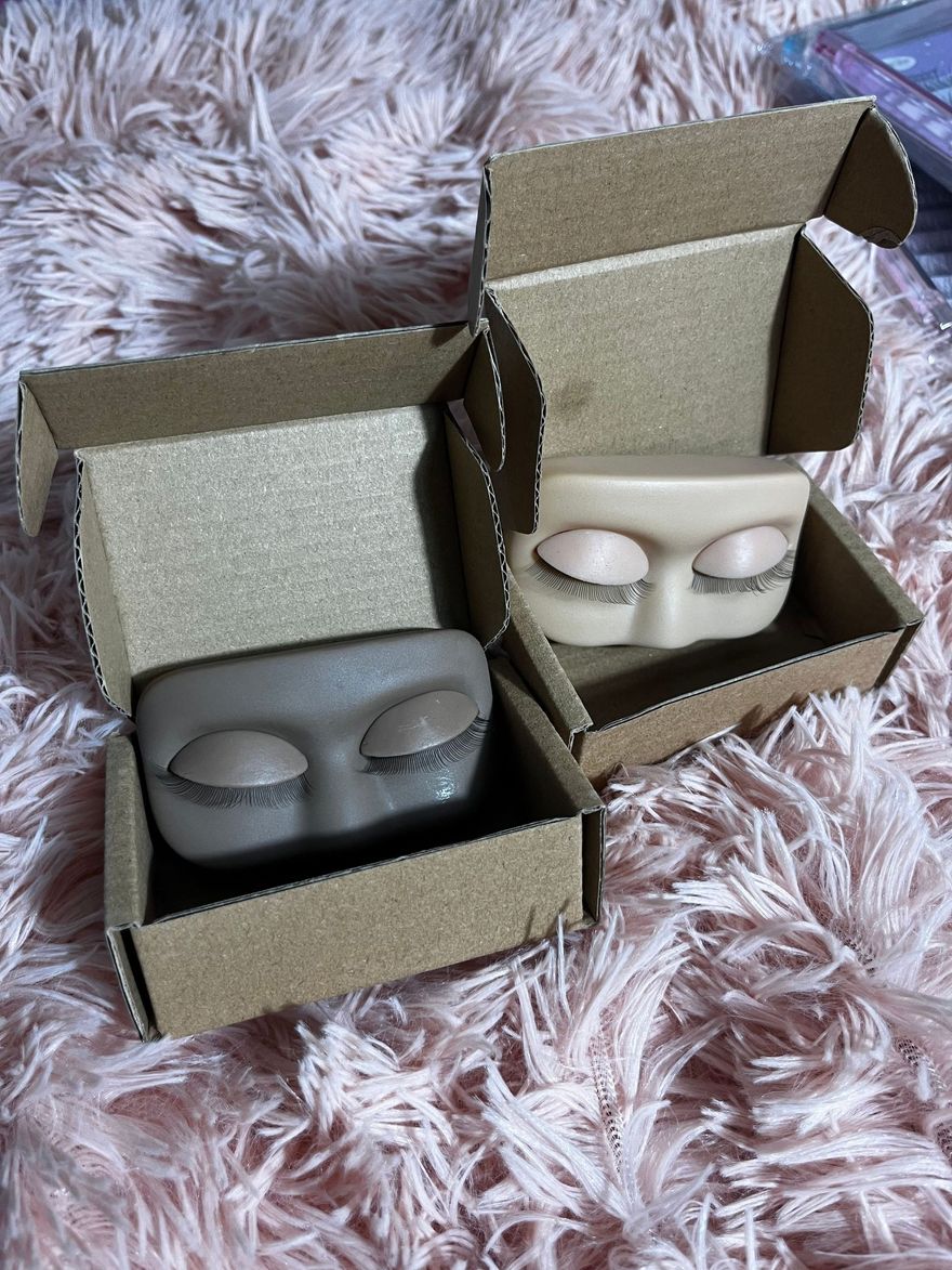 Two face-shaped, closed-eye soap bars inside open cardboard boxes on a pink furry surface. One is gray, one is beige.