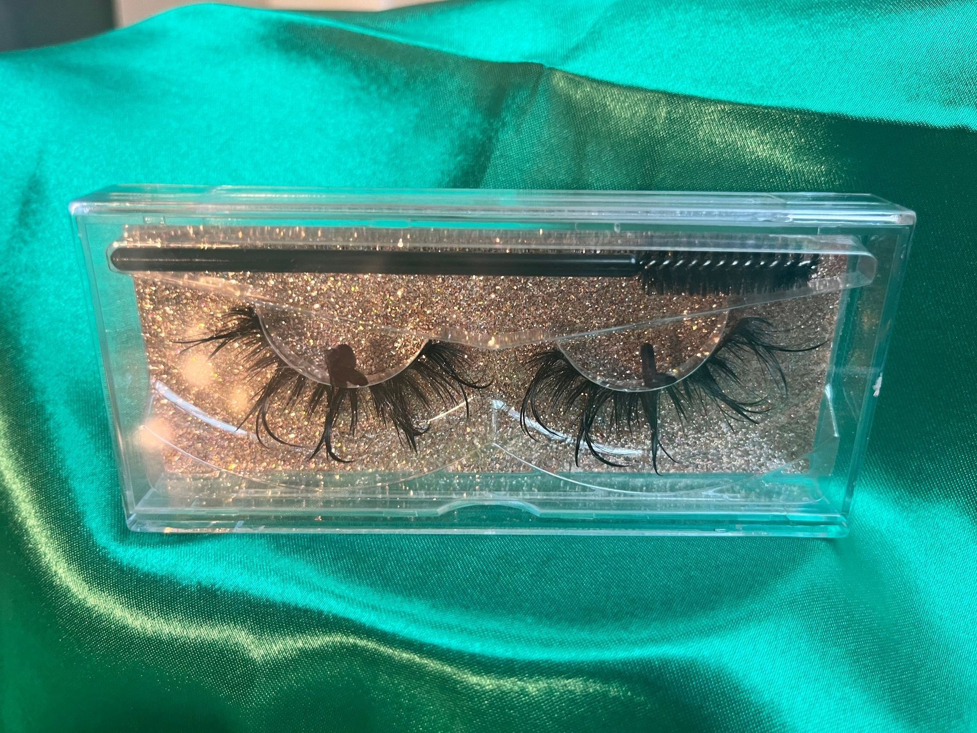Clear plastic case with false eyelashes and mascara wand on gold glitter background.