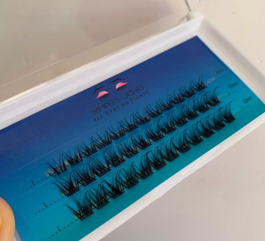 Close-up of a white case holding a tray of black individual eyelash clusters on a blue background.