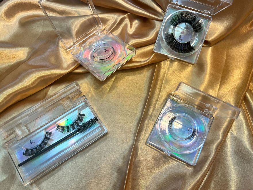Four pairs of false eyelashes in clear plastic cases on a gold satin fabric.