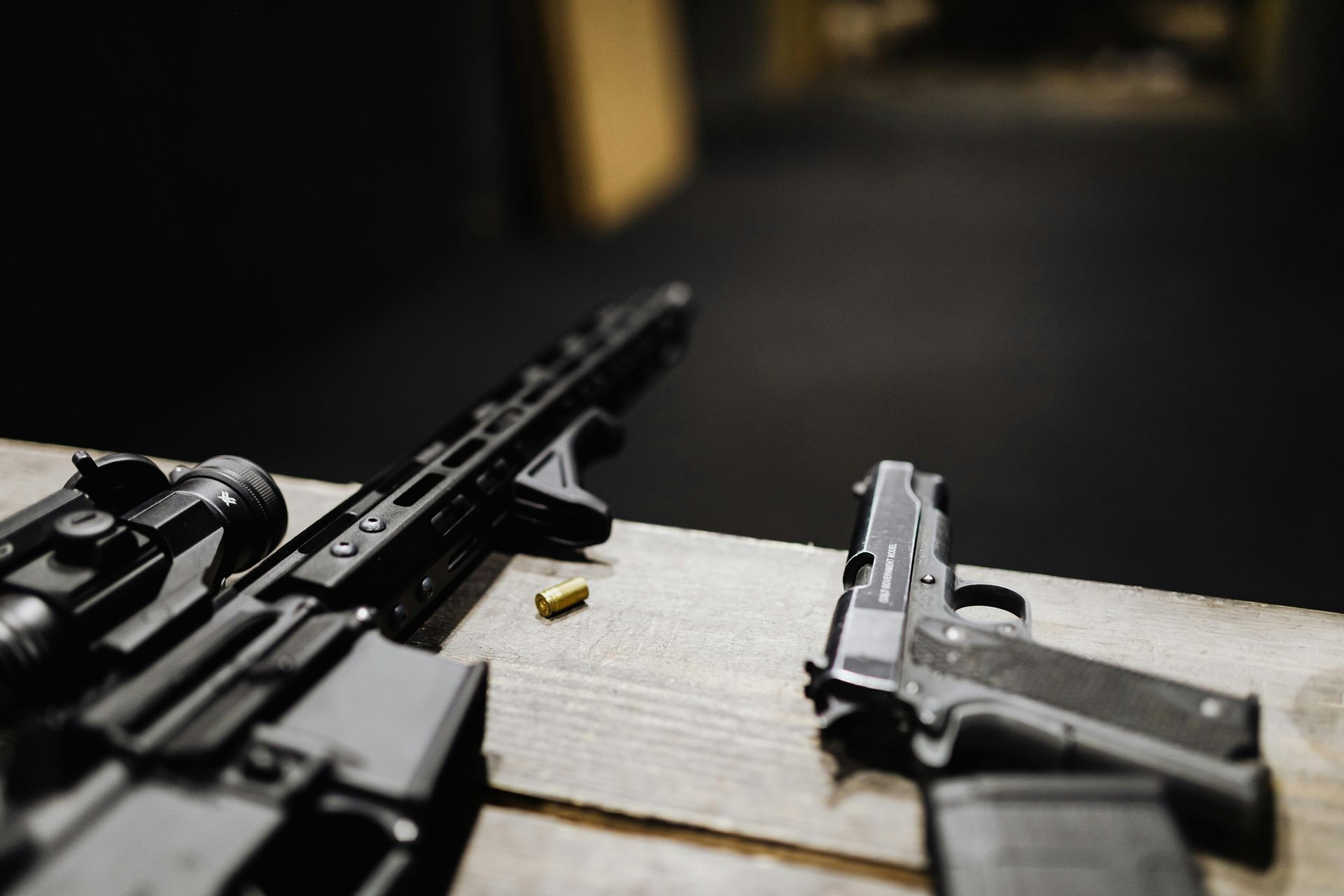 Black rifle and pistol with an ejected bullet on a wooden surface. Dark background.
