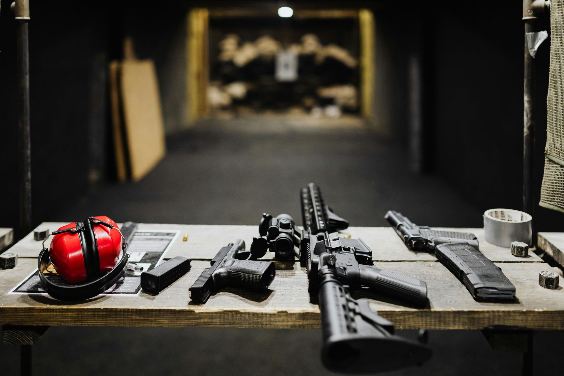 Guns and gear on a shooting range table: red ear protection, pistols, rifle, and targets in the background.