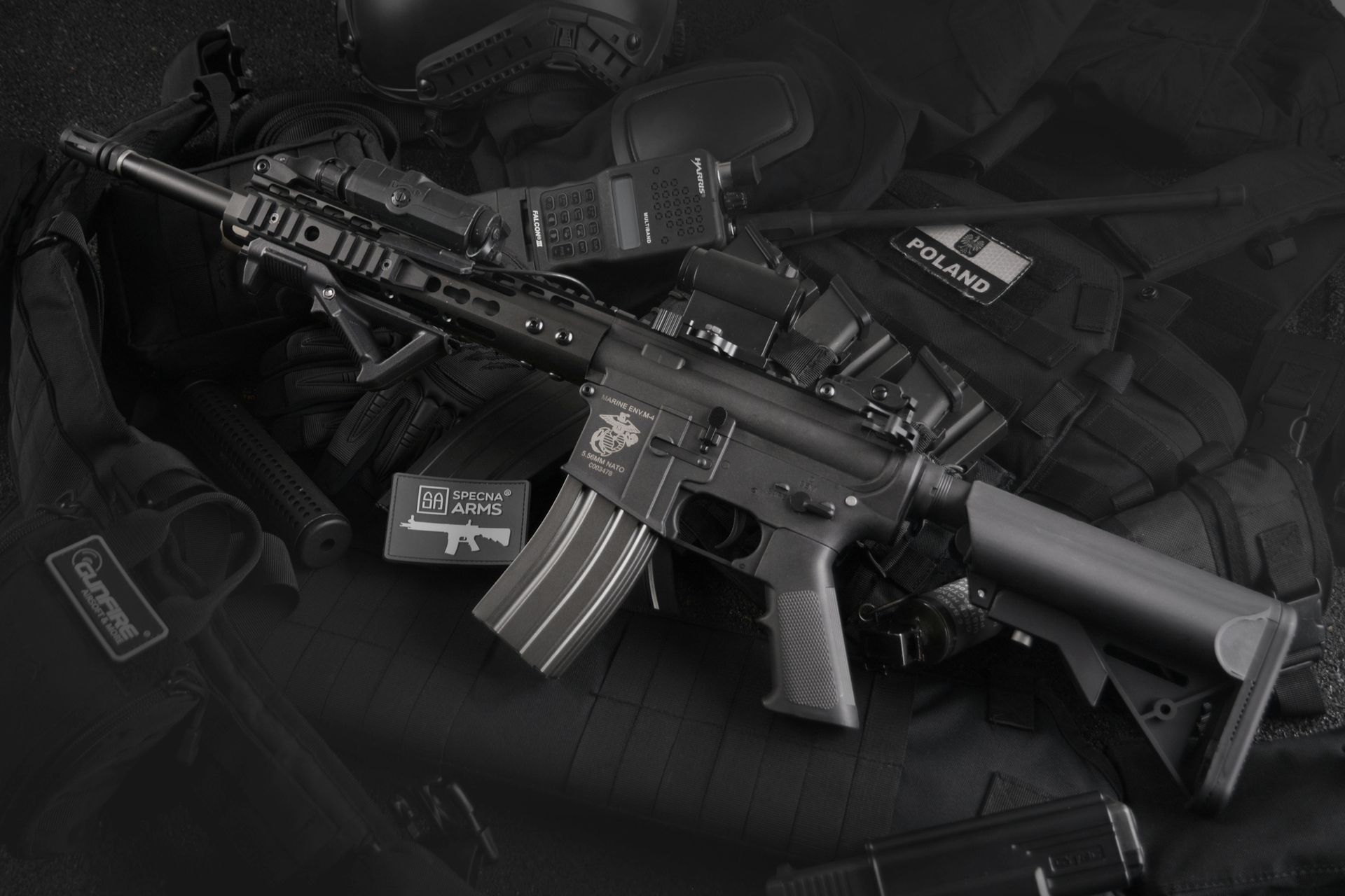 Black and white image of an assault rifle with accessories, resting on a tactical vest and gear.