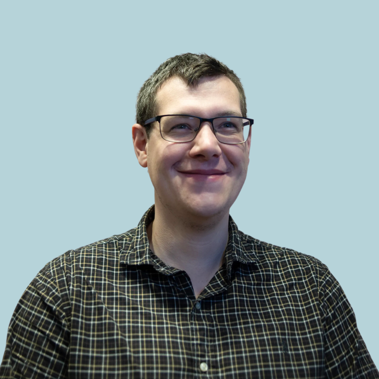 Man with glasses smiles, wearing a black and brown plaid shirt, against a light blue background.