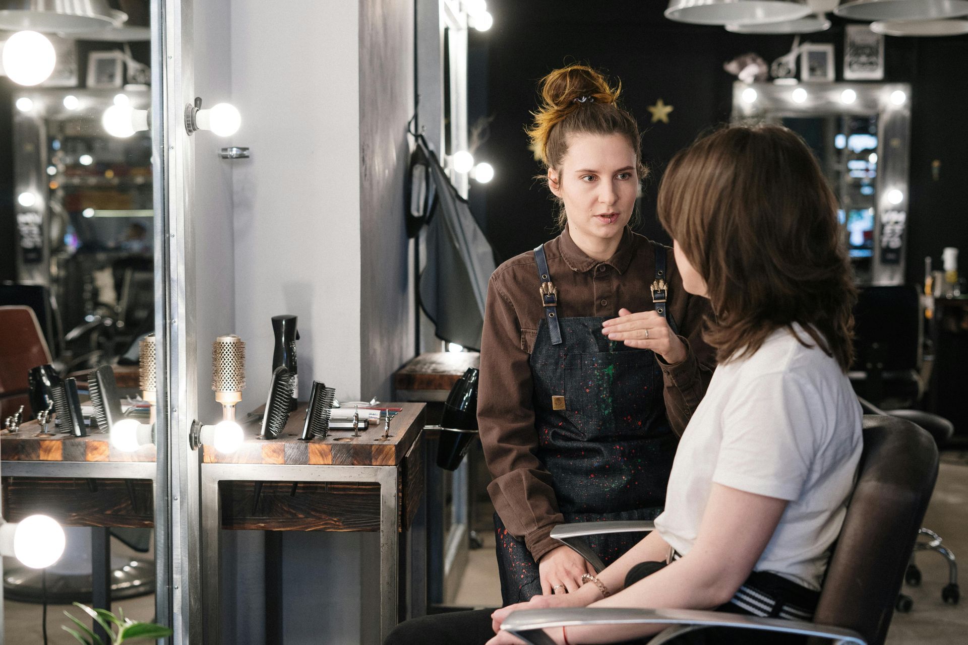 Hair stylist consulting with client in a salon. Stylist gestures while client sits. Mirrors and lights are visible.