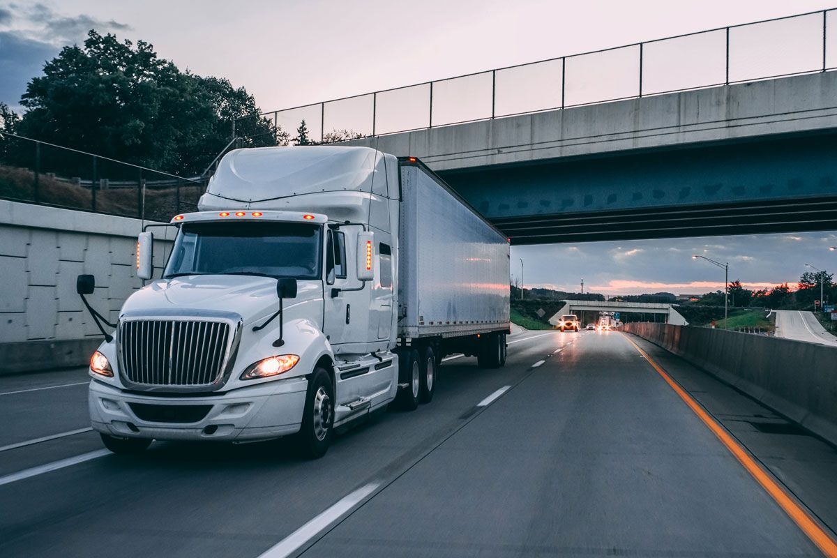 White semi-truck driving on a highway under a bridge at dusk.