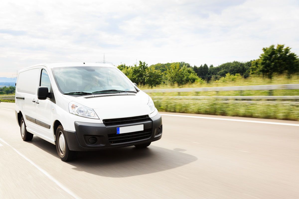 White delivery van driving on a highway, trees in the background, sunny day.