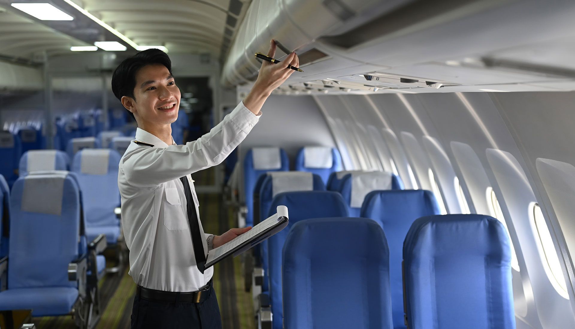 Person in plane cabin checking overhead panel. Smiling. Holding clipboard.