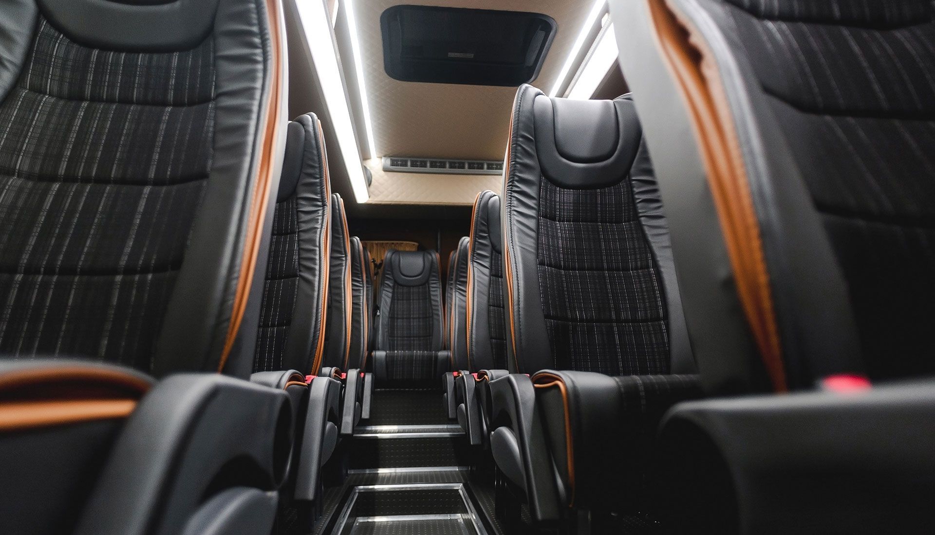 Interior view of a bus with rows of black seats with orange trim, and overhead lights.