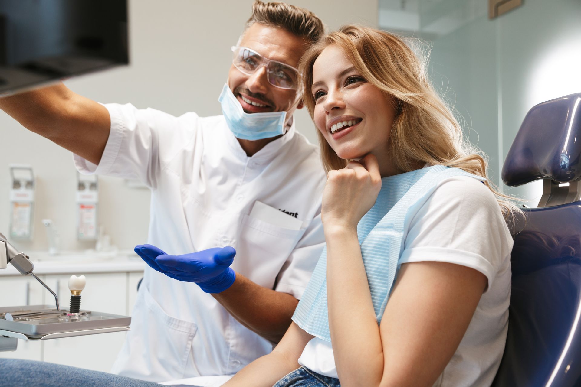 A smiling young woman sits in a medical dentist chair, looking at an x-ray scan, next to a dentist. 